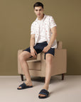 Man sitting on a beige armchair wearing a white patterned shirt, navy shorts, and black sandals against a brown background.