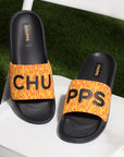 Pair of black CHUPPS sliders with yellow straps featuring red flame patterns and bold black lettering spelling 'CHUPPS', placed on a white chair with green grass background.