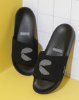 All-black men's sliders with matching sole and white 'C' dot patter resting on a white tile and yellow wall