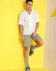 A young man pairs navy blue men’s sliders with grey socks, pista green shorts, and a loose white shirt, styled against a bright yellow background for a bold casual look.