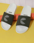 A pair of white and olive green sliders with signature 'C' in dot pattern on a yellow floor resting on orange rolled up chart paper.