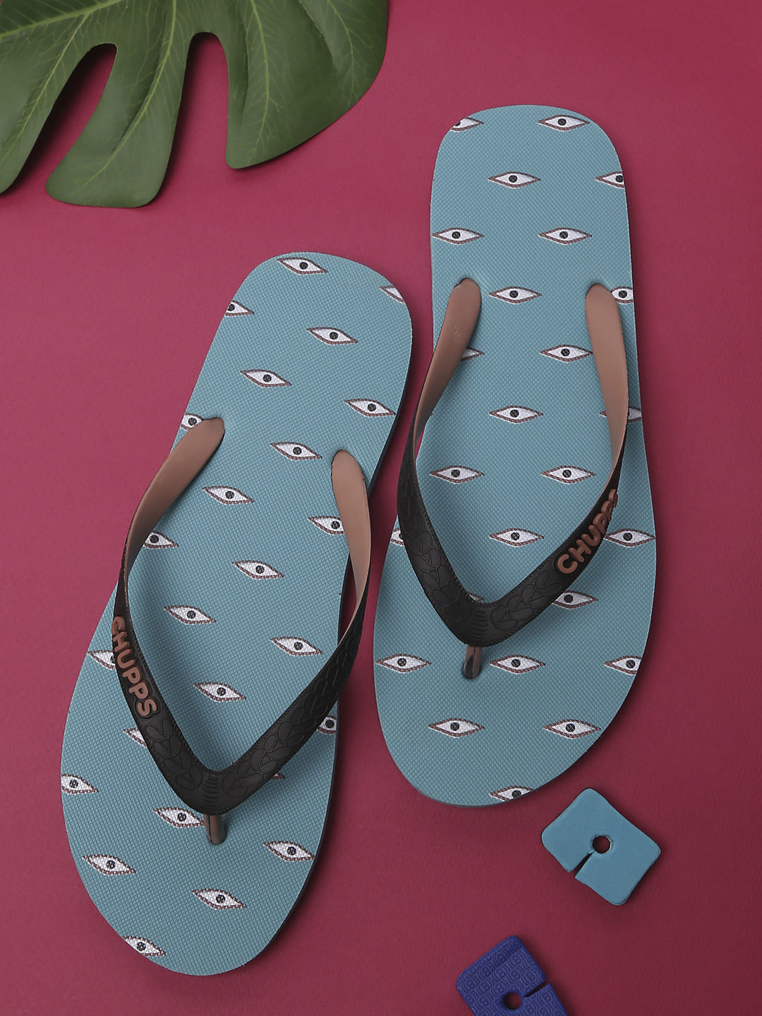 Chupps turquoise flip-flops with quirky eye print and black straps, stylish men’s summer slippers on a vibrant pink background.