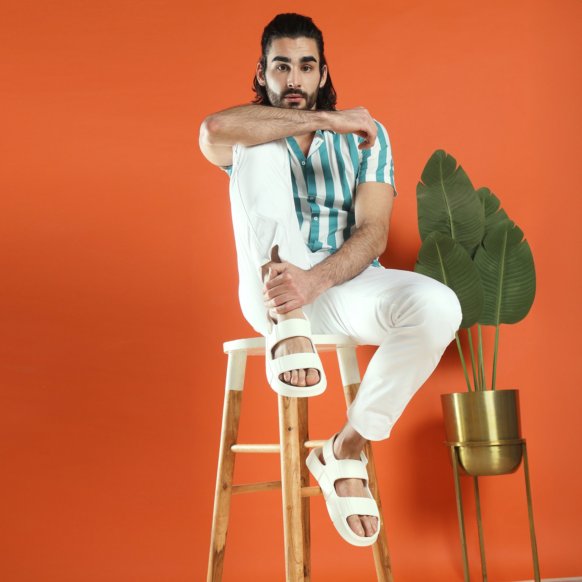 Chunky Sandals_almond_white chucky man sitting on a wooden stool against an orange background with a plant to his right.
