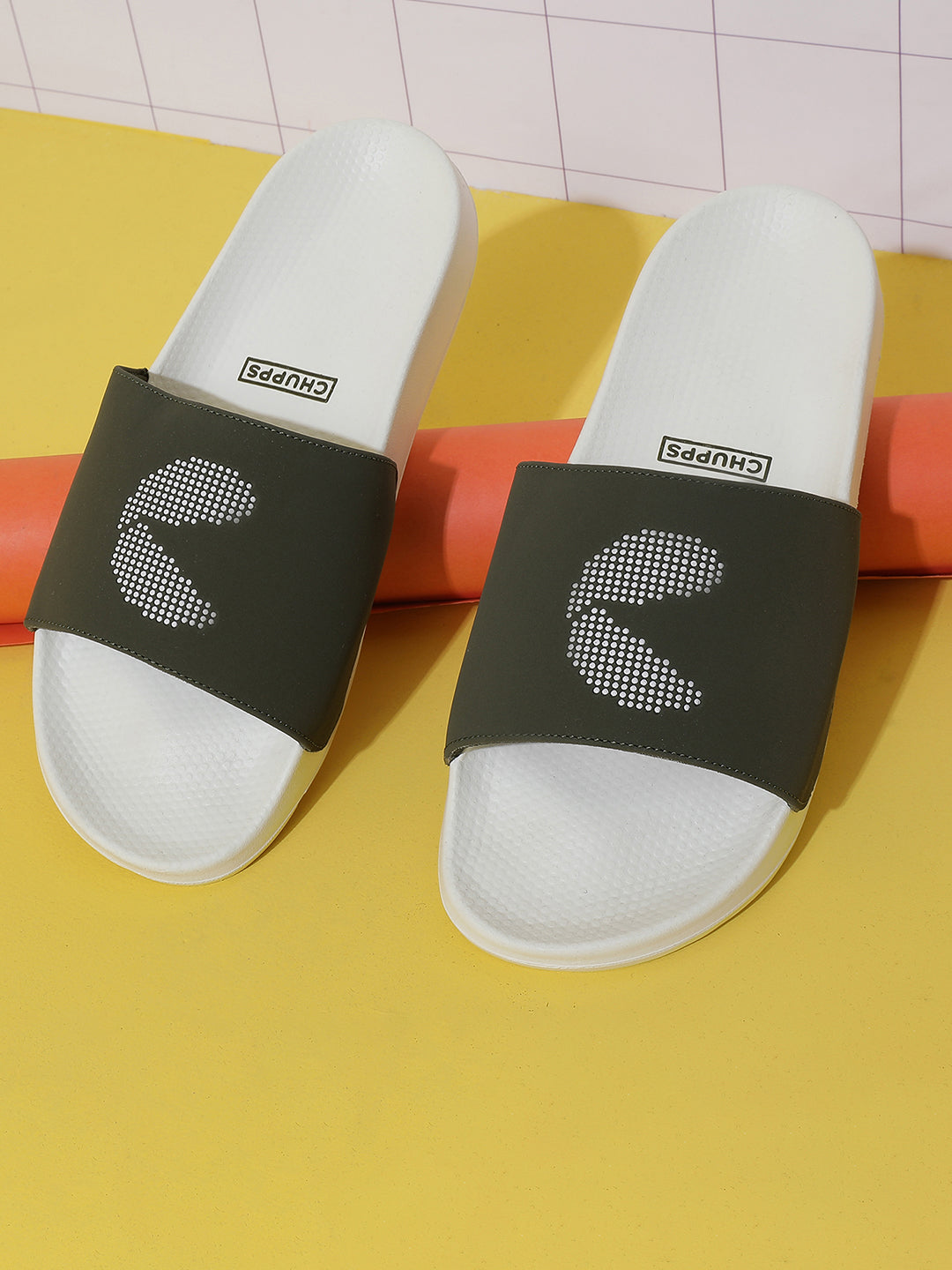 A pair of white and olive green sliders with signature 'C' in dot pattern on a yellow floor resting on orange rolled up chart paper.